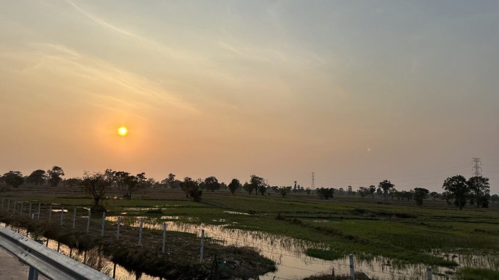 Ein stimmungsvoller Sonnenuntergang über grünen Feldern, Bäumen und Wasserflächen, dazu ein Zaun samt Straße am linken Rand.