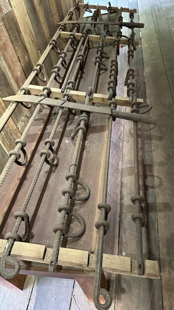 Hier siehst du eine alte Vorrichtung aus Holz und Metall mit Stangen und Ösen auf einem Holztisch vor rustikaler Holzwand.