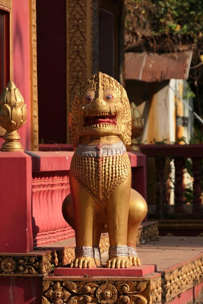Vor einem gold-roten Tempel wacht eine kunstvoll gestaltete goldene Löwenstatue, umgeben von Grün und weiteren Gebäuden.