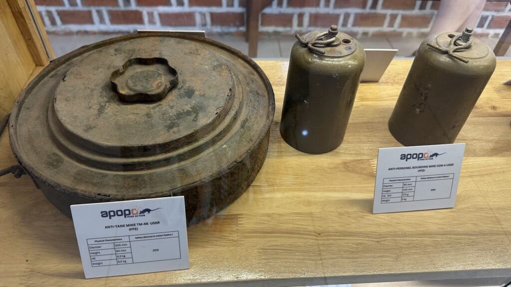 In einer Vitrine liegen eine große, rostige Panzerabwehrmine und zwei grüne Antipersonenminen neben Infokarten mit APOPO-Logo.