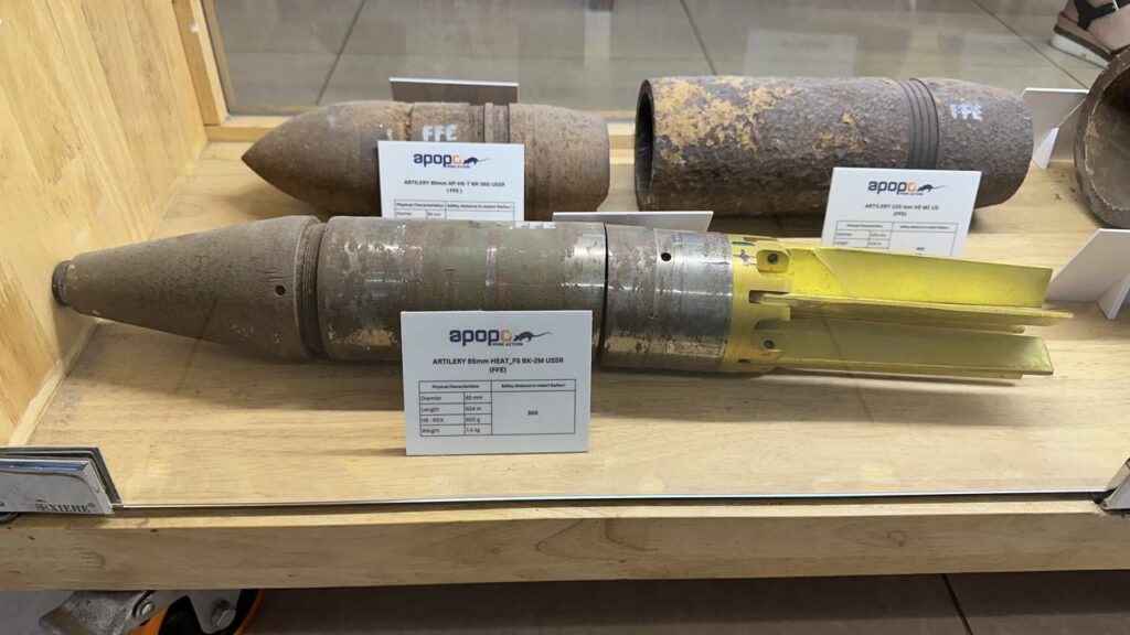 In einer Glasvitrine auf Holz liegen drei alte, rostige Artilleriegranaten mit Infokarten - die vordere hat einen gelben Leitflügel.