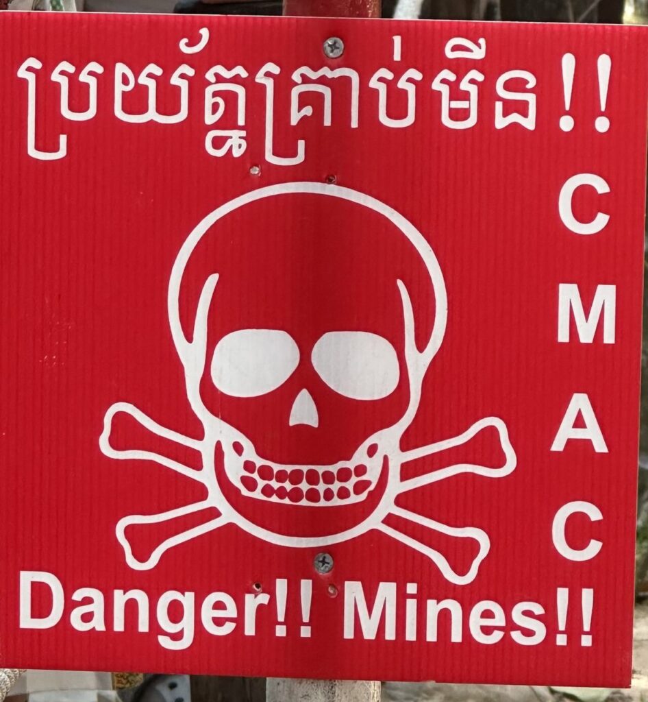Ein rotes Warnschild mit Totenkopf, "Danger!!! Minen!!", Text auf Khmer und "CMAC" seitlich - Achtung, Landminen!.