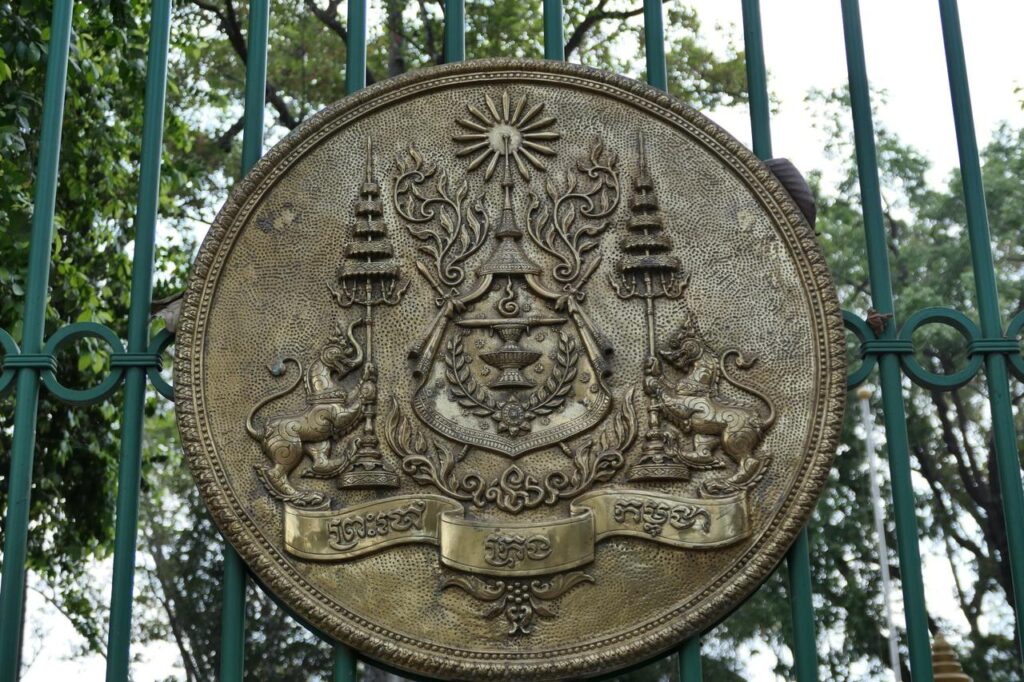 Ein kunstvoll verziertes Bronze-Emblem mit Fabelwesen und Khmer-Schrift auf grünem Zaun, umgeben von Bäumen im Hintergrund.