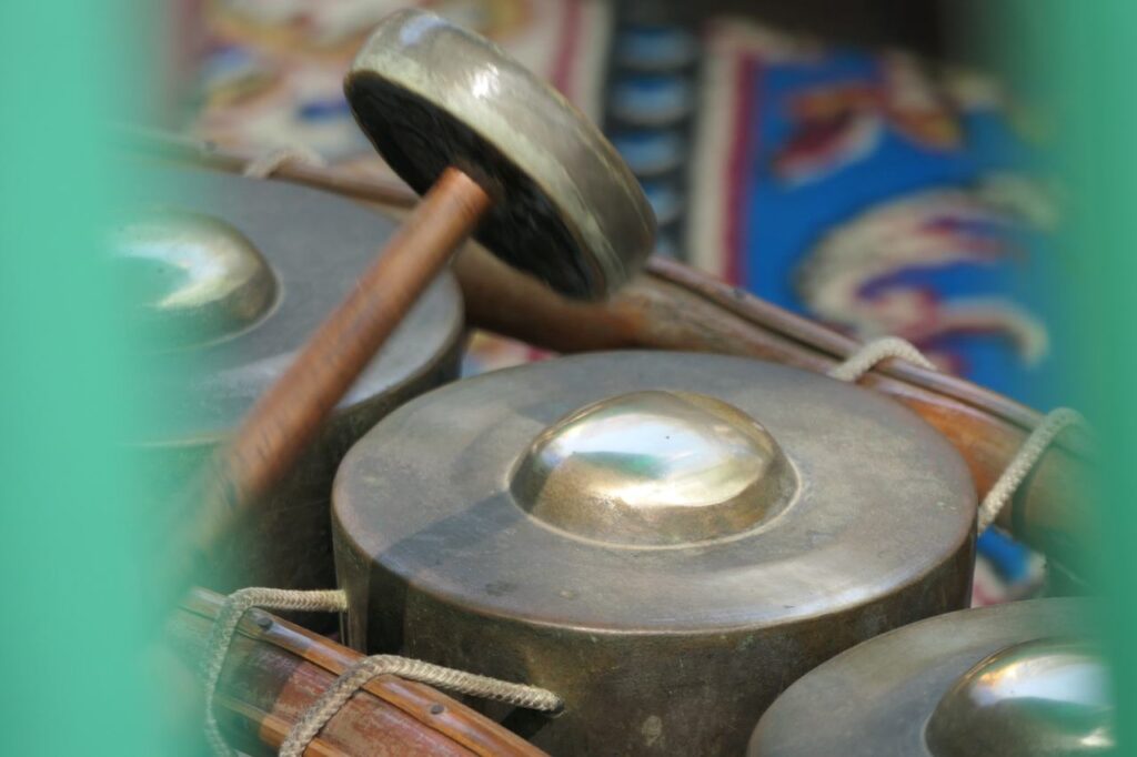 Detailreiche Nahaufnahme eines indonesischen Gamelan-Instruments mit Bronzeklangschalen, Schlegel und buntem Stoff im Hintergrund.
