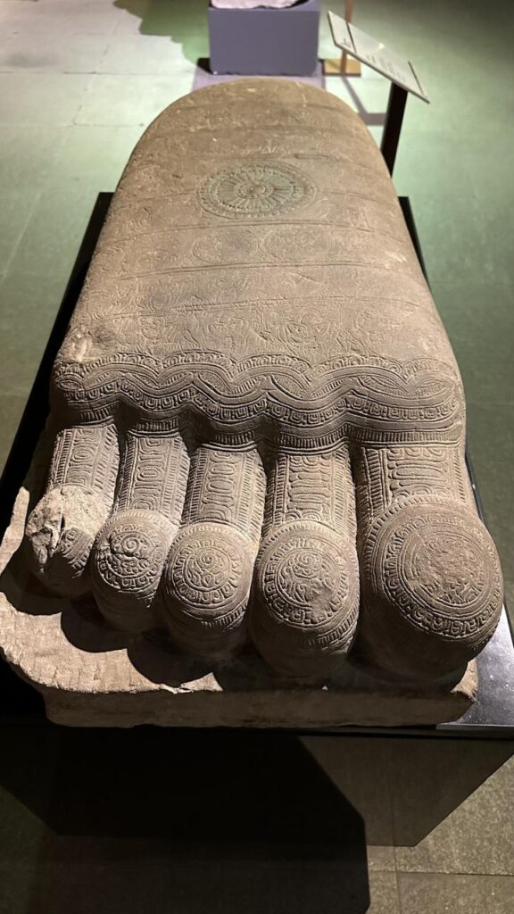 Im Museum bewundern wir einen kunstvoll verzierten steinernen Fuß, inspiriert vom Königreich der Khmer, auf schwarzem Sockel.