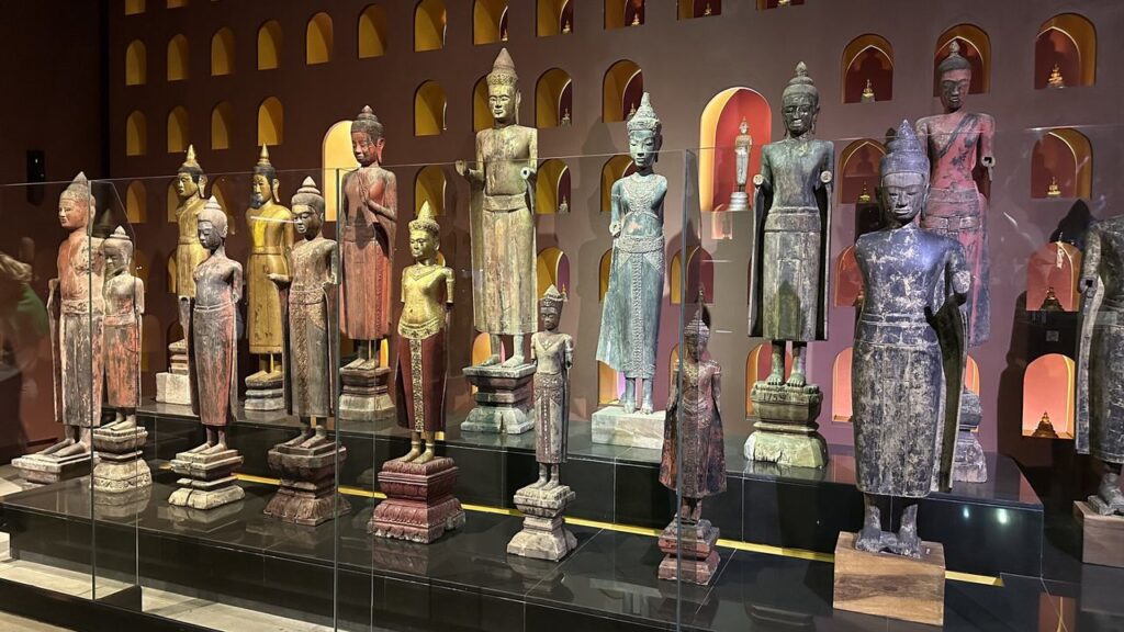 Verschiedene buddhistische Statuen in bunten Farben und Größen stehen und sitzen ordentlich im Museum vor einer dekorativen Wand.