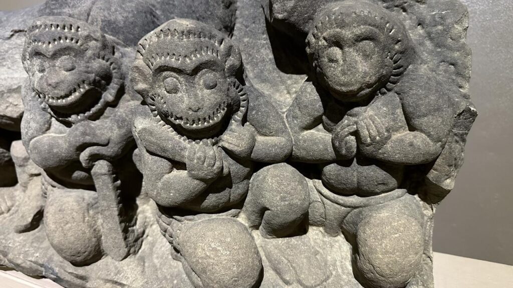 Drei kunstvoll gemeißelte Stein-Affen aus dem Khmer-Königreich stehen lebhaft nebeneinander vor rauem Fels und halten Gegenstände.