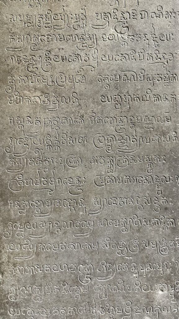 Eine alte Steintafel mit kunstvoll eingravierten, geschwungenen Schriftzeichen aus Süd- oder Südostasien auf grauem Stein.