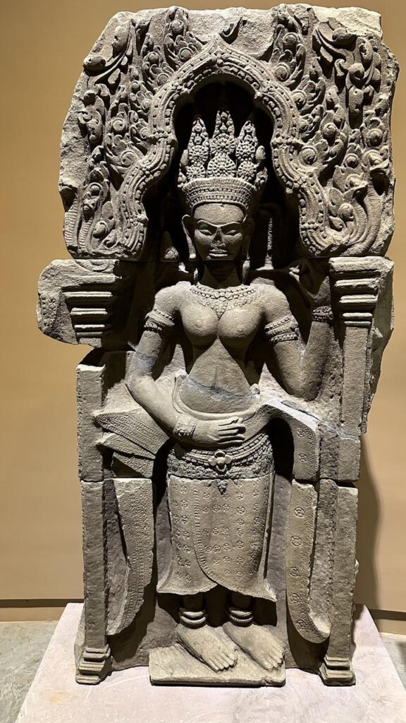 Eine kunstvolle Khmer-Skulptur zeigt eine Göttin mit edler Krone und Schmuck, elegant stehend im blumig verzierten Steinbogen.
