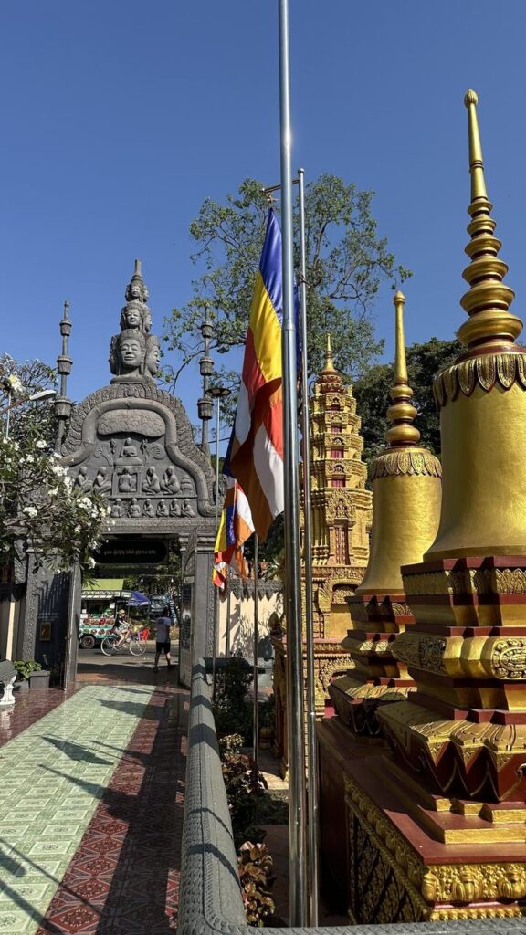 Bunte Flaggen und goldene Stupas schmücken das Tor eines Khmer-Tempels, strahlend im Sonnenschein vor blauem Himmel und Bäumen.