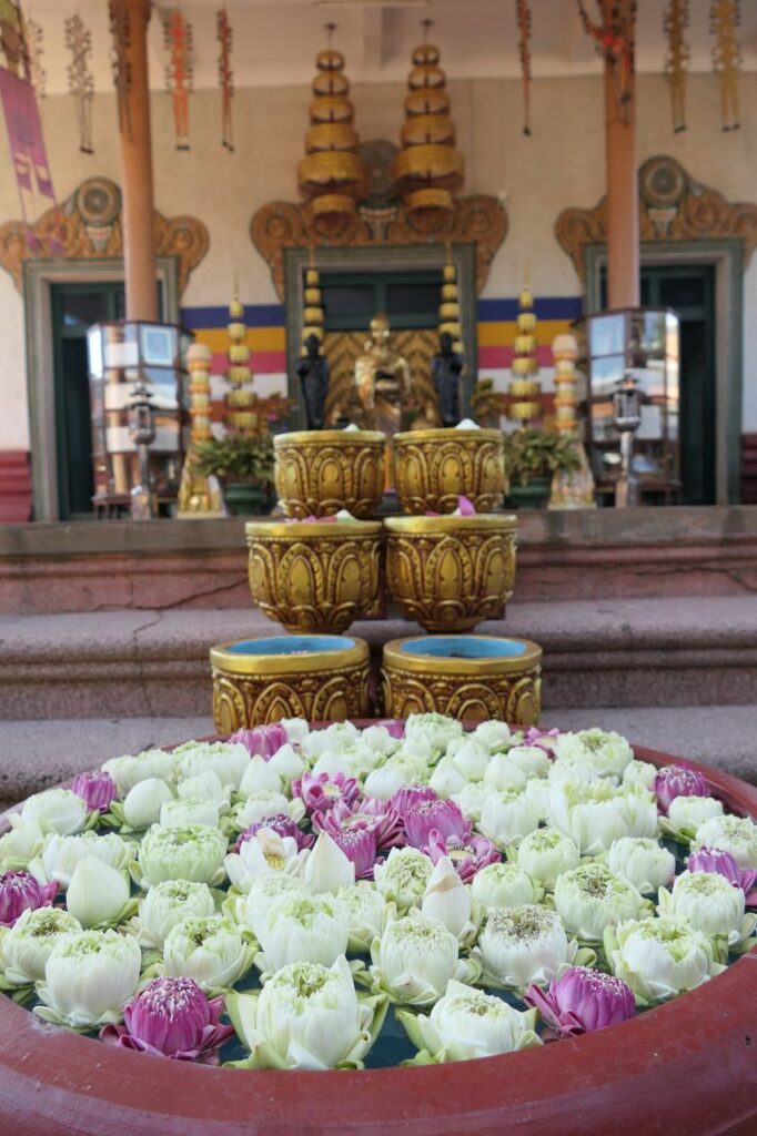 Eine große Schale mit schwimmenden weißen und rosa Lotusblüten steht vor goldenen Trommeln und einem reich geschmückten Altar.