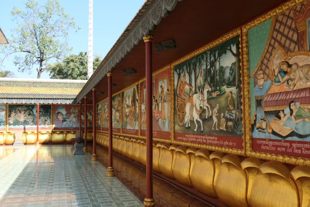 Ein Tempelflur mit farbenfrohen Wandmalereien zu buddhistischen Geschichten, verziert mit goldenen Lotus-Ornamenten am Sockel.