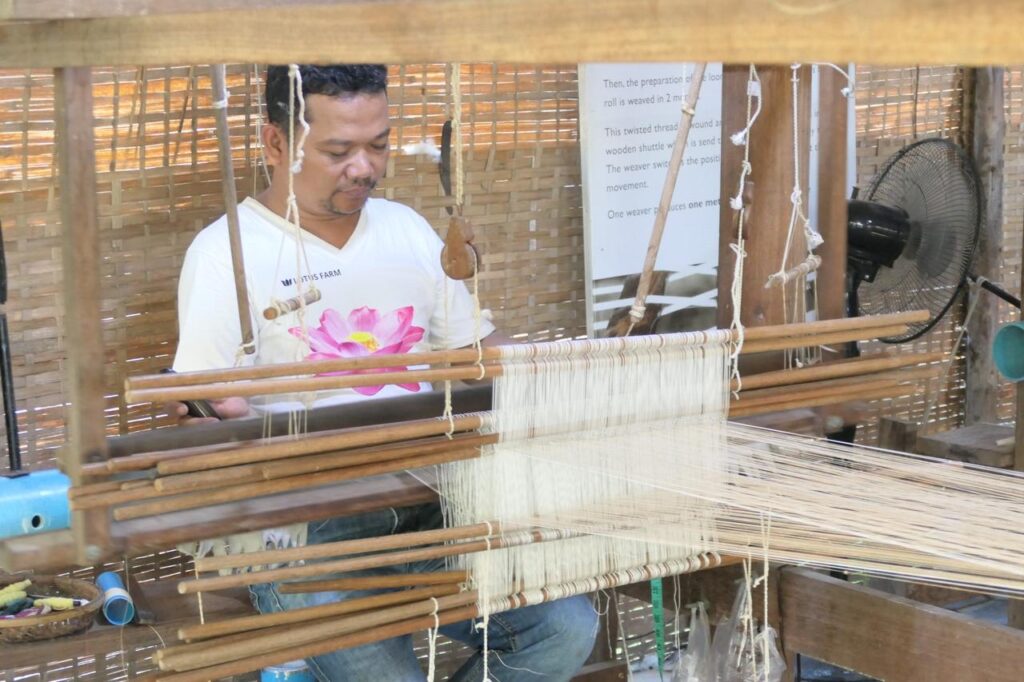 Ein Mann webt mit Hingabe weiße Fäden am traditionellen Webstuhl vor Bambuswänden - ein Stück Khmer-Tradition live erleben.