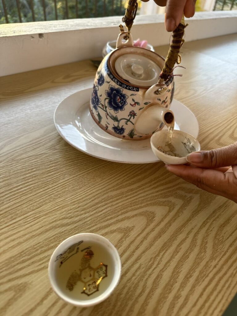 Eine Hand gießt Tee aus einer hübschen, blumigen Teekanne in eine Tasse; daneben steht noch eine weitere auf Holztisch am Fenster.