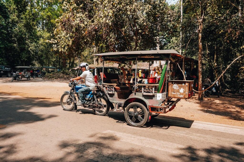 Auf einer sonnigen Straße im Königreich der Khmer fährt jemand fröhlich mit einem Tuk-Tuk durch viel Grün.