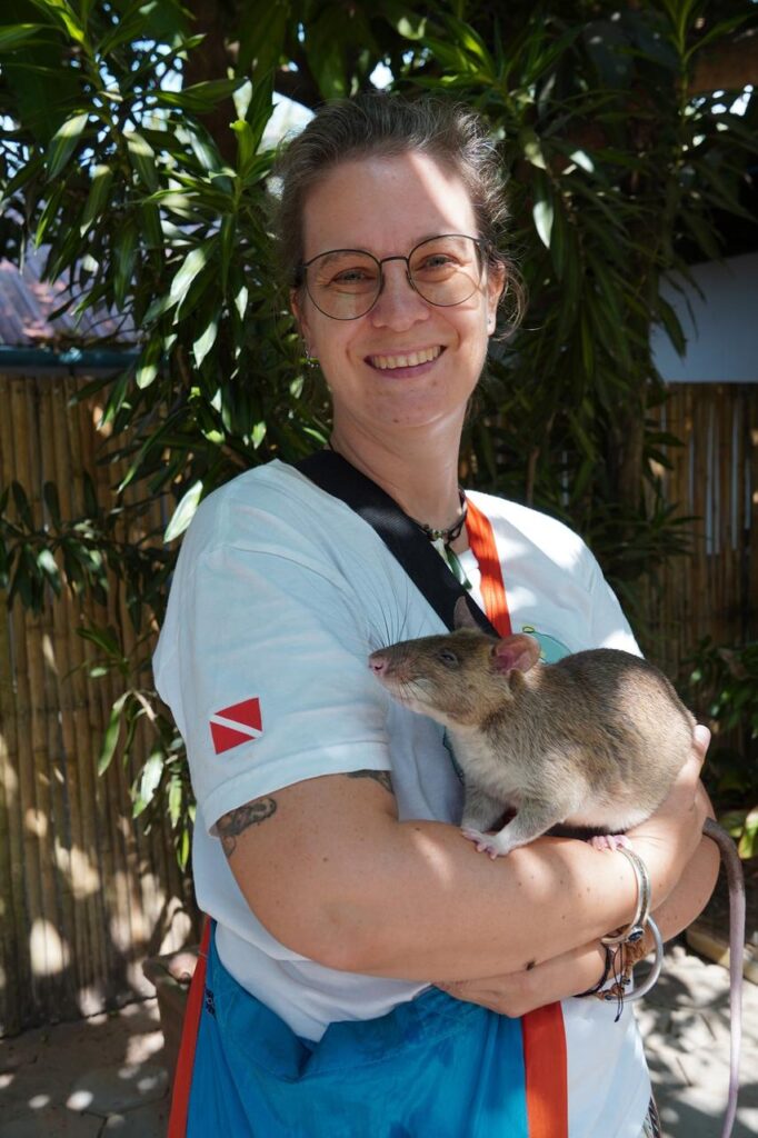 Eine Person mit Brille und weißem Hemd lächelt, hält eine große Ratte im Arm und steht vor grünen Pflanzen und Bambuszaun.