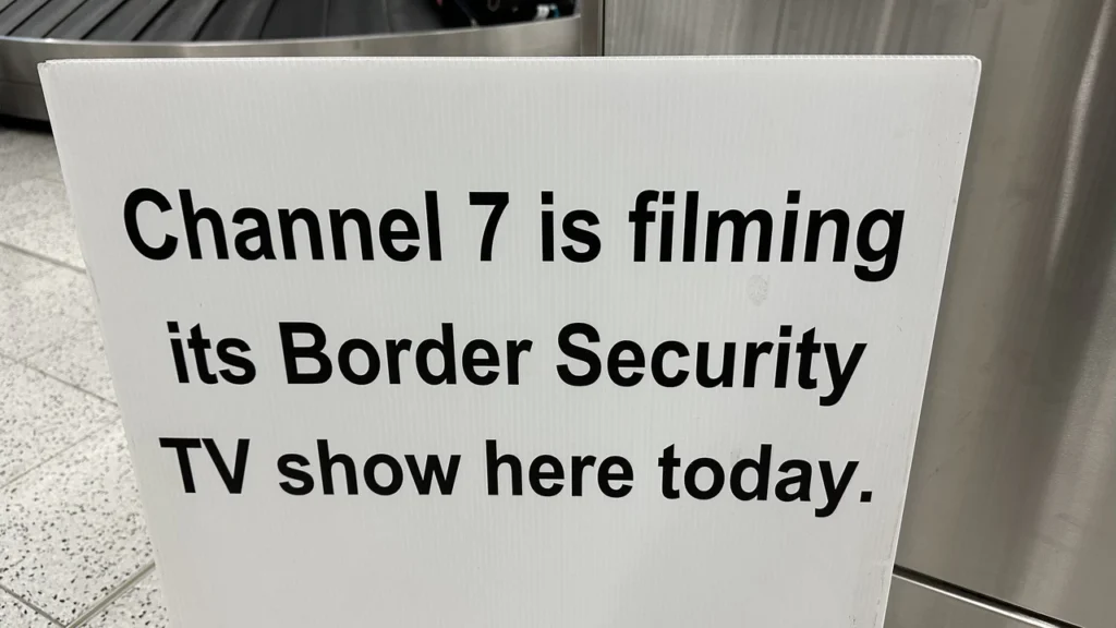 Ein weißes Schild mit schwarzer Aufschrift informiert freundlich, dass Channel 7 heute die Sendung "Border Security" dreht - vermutlich am Flughafen.