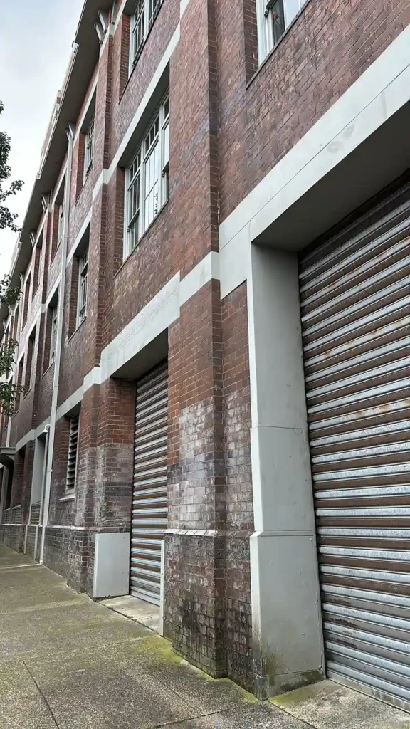 Ein hohes Backsteingebäude mit großen, geschlossenen Rolltoren und vielen Fenstern. Der Gehweg verläuft direkt daneben.