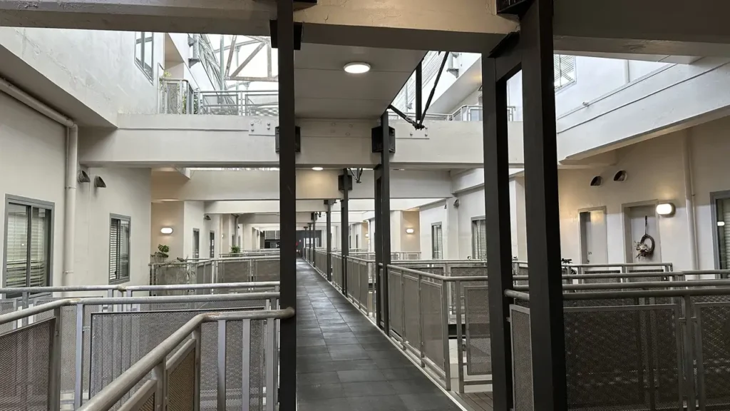 Ein heller Flur in einem modernen Apartmenthaus mit Metallgeländern, grauem Boden und Türen, Fenster und Balkone auf beiden Seiten.