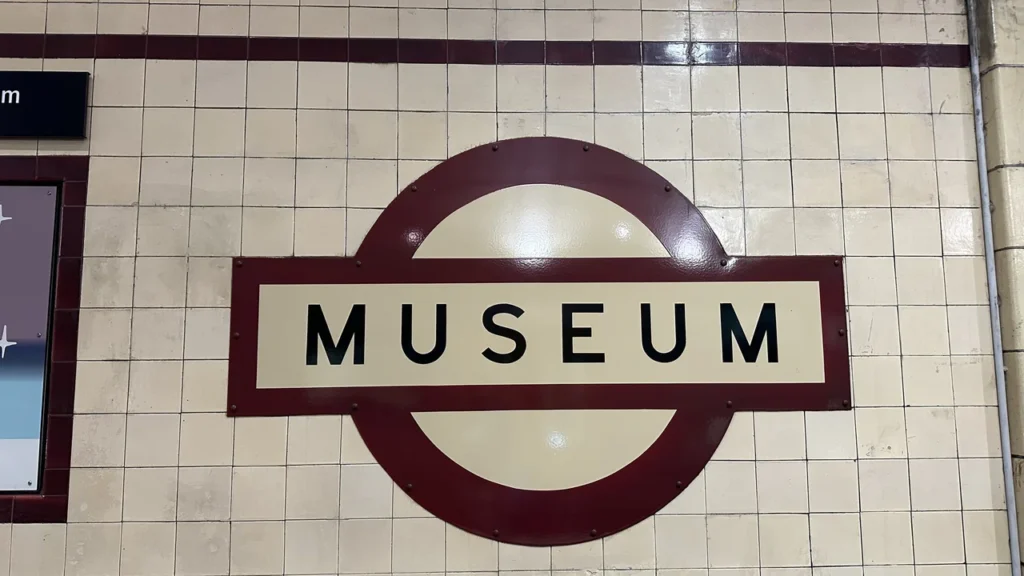 Ein Museums-Schild mit schwarzer Schrift auf cremefarbenem Grund und rotem Rand schmückt freundlich eine geflieste Wand.