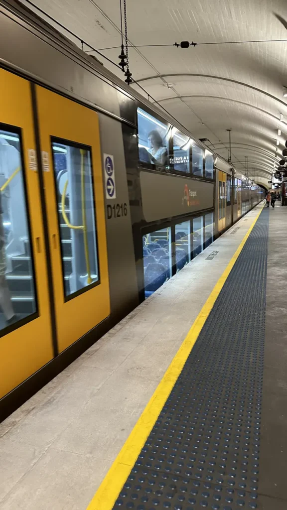 Ein gelb-silberner Zug hält in einer modernen U-Bahn-Station mit weiß geschwungener Decke, wenige Fahrgäste warten am Bahnsteig.