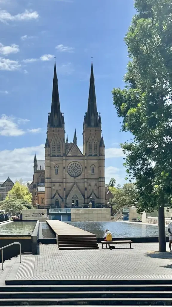 Im Hintergrund ragt eine große Kathedrale mit zwei Türmen auf, rechts ein Baum. Vorne sitzt jemand am Wasser und ein Weg führt hinüber.