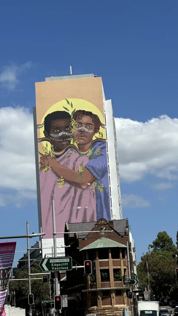 An einer hohen Hauswand strahlt ein buntes Wandbild: Zwei dunkelhäutige Kinder, nah beieinander, mit Blumen im Gesicht. Darunter Ampeln.