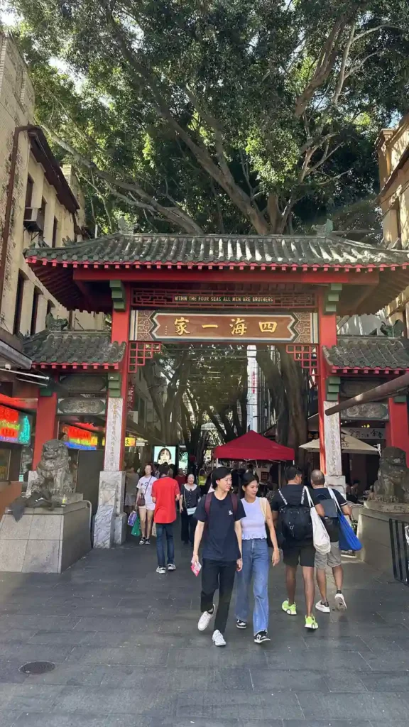 Unter einem rot-goldenen Chinatown-Tor schlängeln Menschen vorbei an Löwenstatuen, umgeben von Bäumen und kleinen Läden.