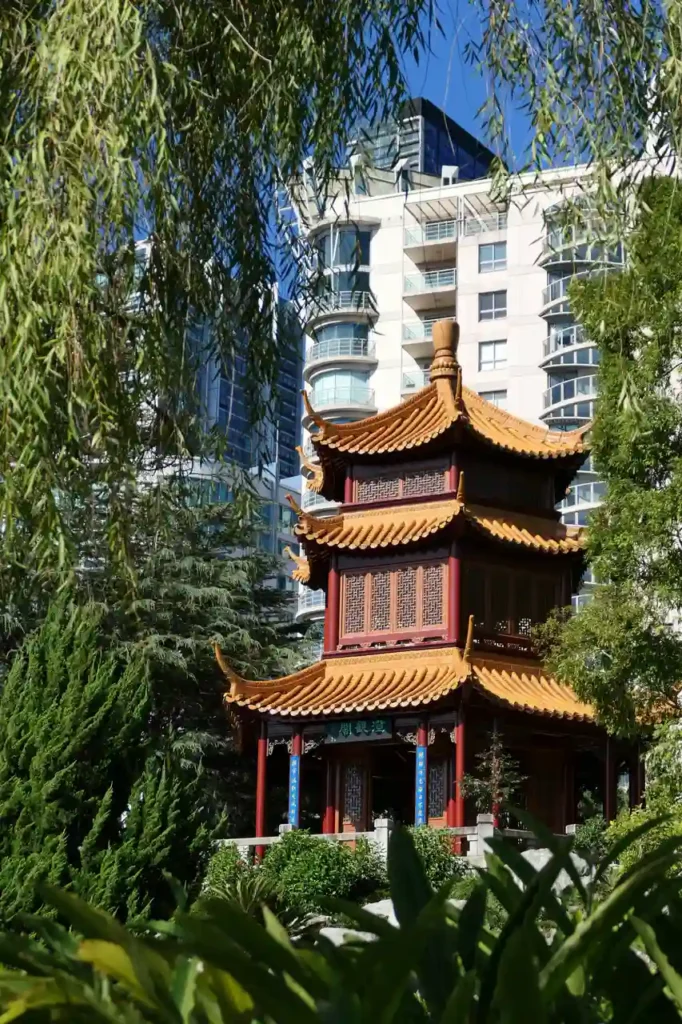 Ein chinesischer Pavillon mit goldenen Dächern liegt idyllisch im Grünen, umgeben von modernen Hochhäusern und blauem Himmel.