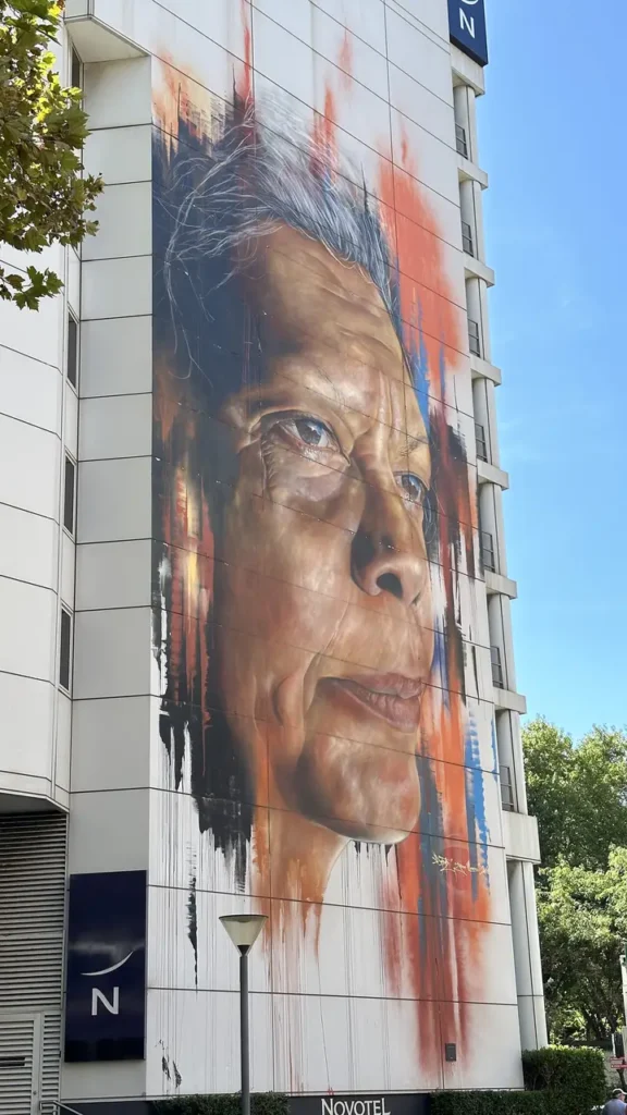 Ein großes Wandbild zeigt das realistische Porträt einer älteren Person mit grauen Haaren an einem weißen Gebäude, Bäume und Novotel-Schild im Blick.