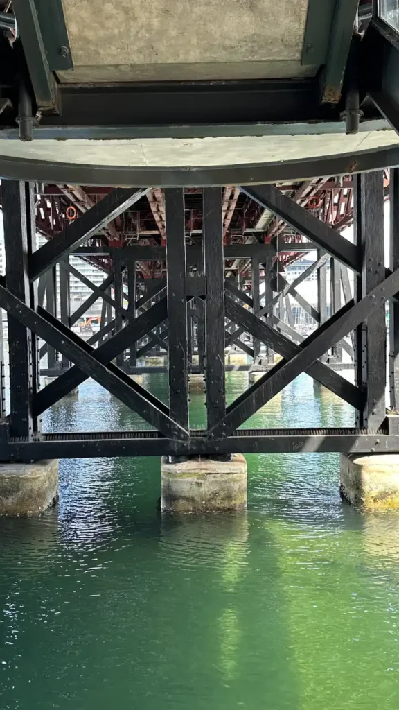 Ein Blick unter eine Brücke: Holz- und Metallstreben kreuzen sich über grünlichem Wasser, Sonnenlicht spielt auf der Oberfläche.