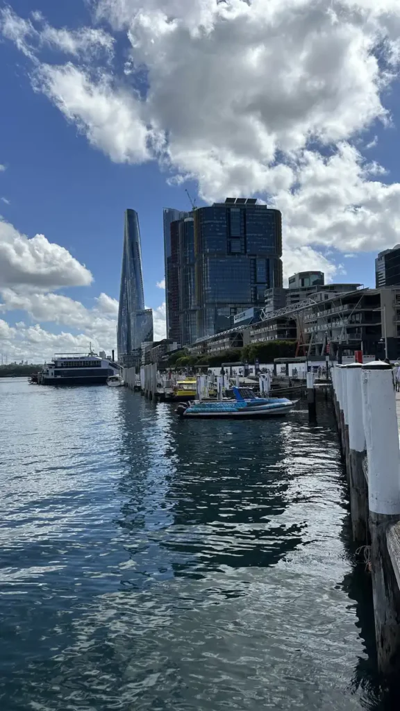 Am Wasser liegen Boote an einem Holzsteg, dahinter moderne Hochhäuser. Das ruhige Wasser spiegelt den teils wolkigen Himmel.