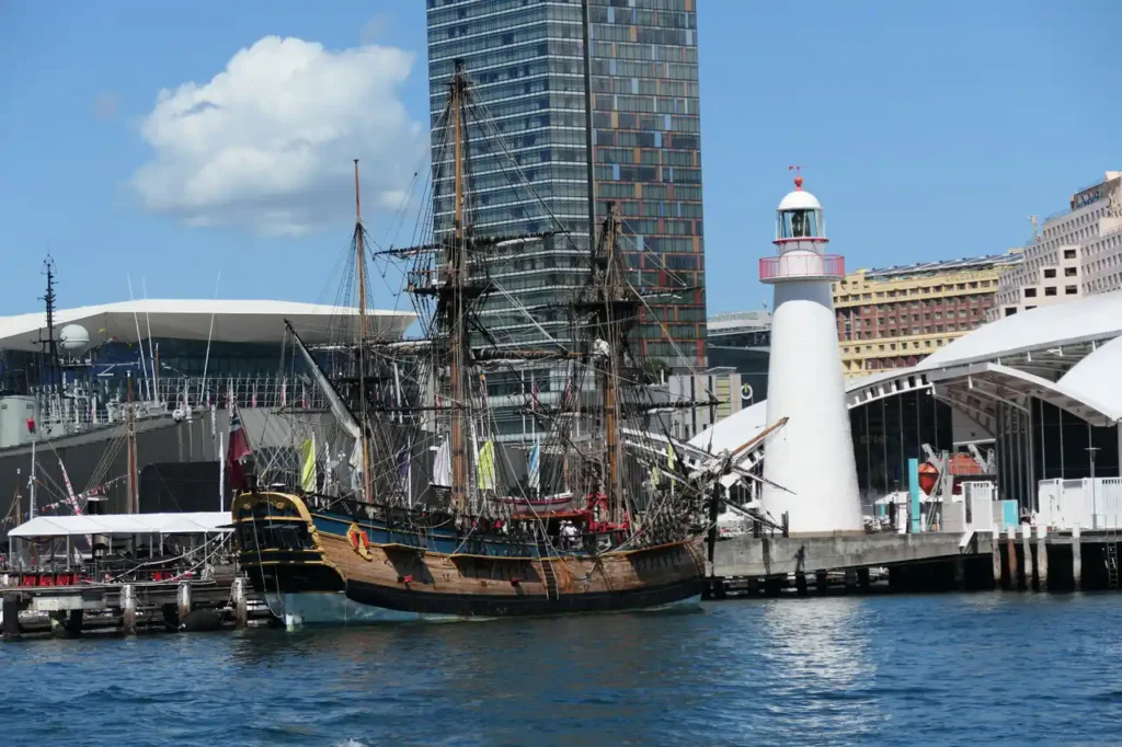 Ein altes Segelschiff liegt im modernen Hafen, nahe eines weißen Leuchtturms. Im Hintergrund Glasbauten und blauer Himmel.