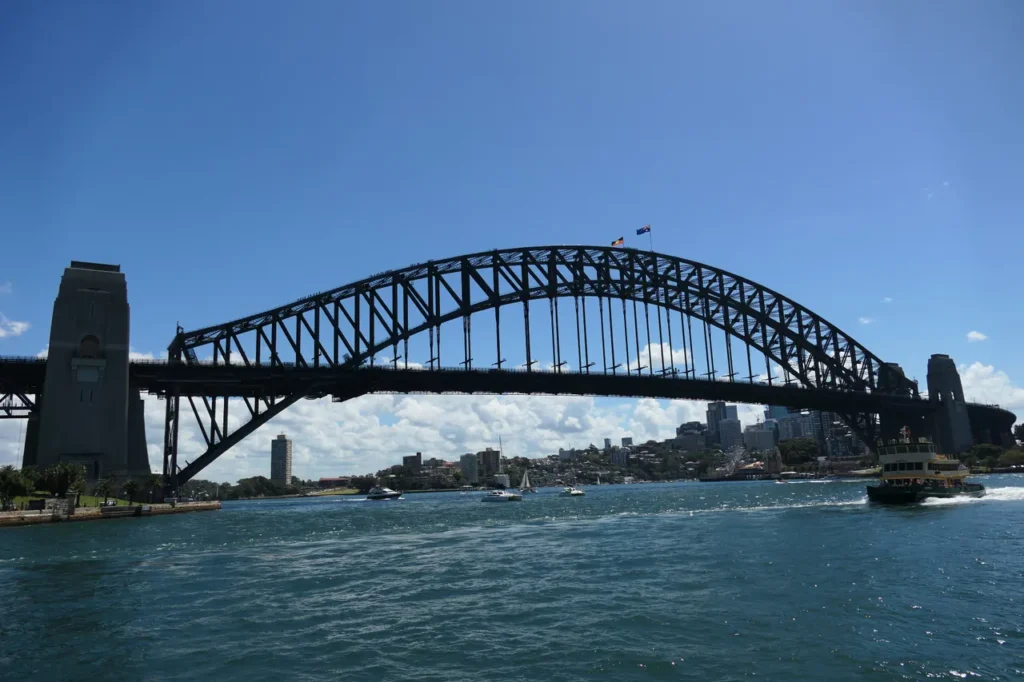 Die Sydney Harbour Bridge erstreckt sich über das blaue Wasser, Boote und eine Fähre sind zu sehen, dahinter die Skyline der Stadt.