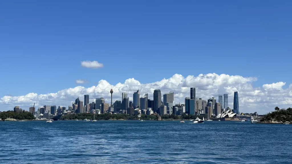 Blick über den Hafen auf Sydneys Skyline mit Opernhaus, hohem Tower und entspannten Booten unter freundlichem Himmel.