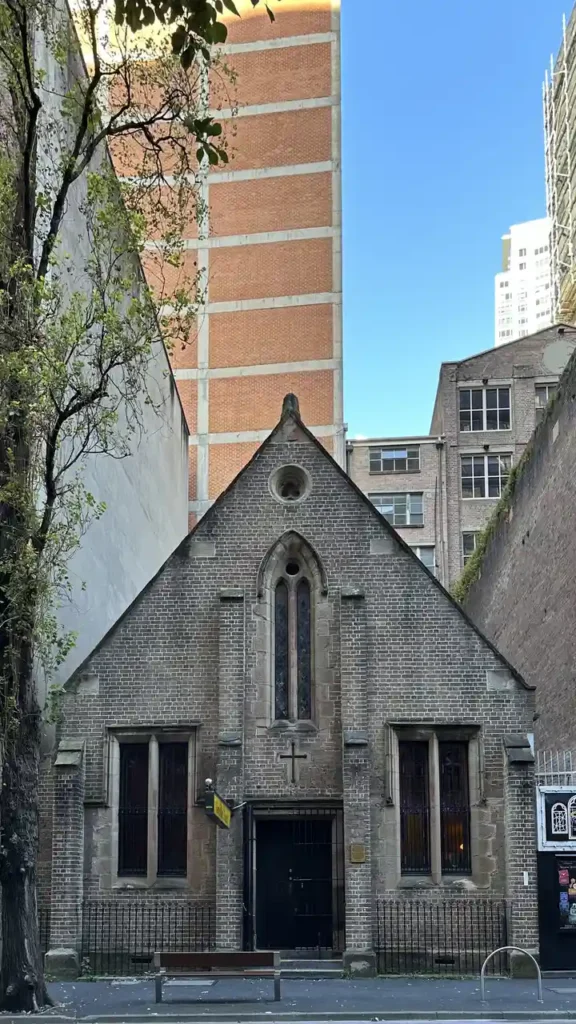 Zwischen hohen, modernen Gebäuden steht eine kleine, alte Backsteinkirche mit Spitzdach, Rundbogenfensterstern und Kreuz. Links ein Baum.