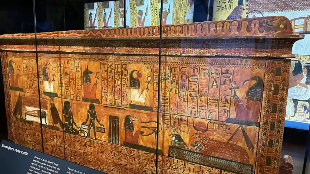 In einem Museum bewundern wir einen altägyptischen Sarkophag, bunt bemalt mit Göttern wie Anubis und Osiris, goldener Hintergrund.