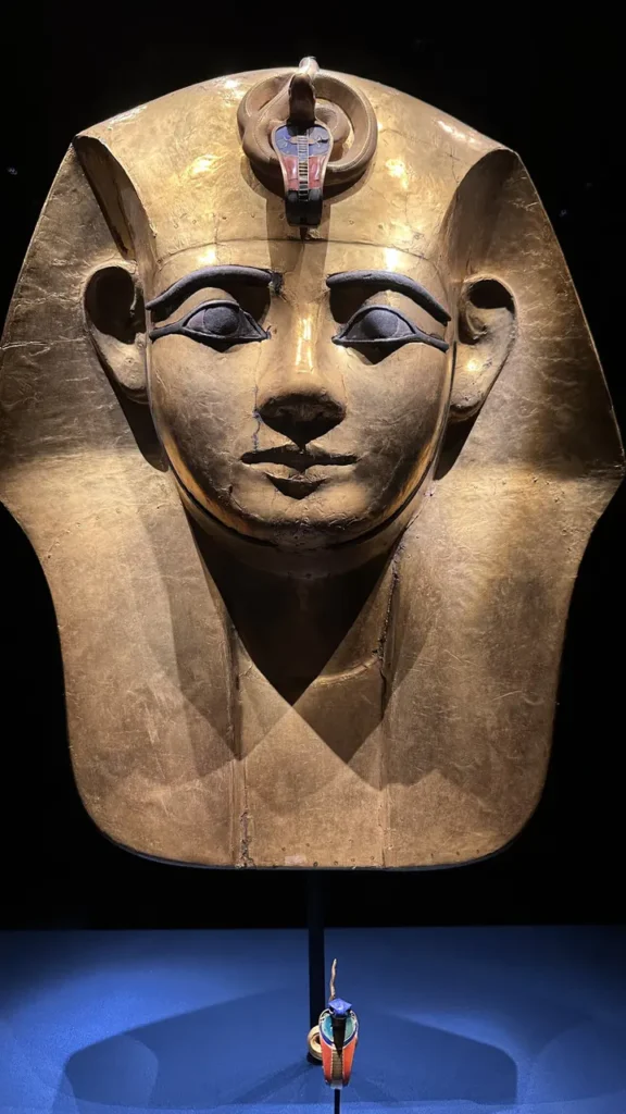 Eine goldene Pharaomaske mit blauer und schwarzer Augenpartie, Uraeus-Kobra und buntem Objekt davor vor dunklem Hintergrund.