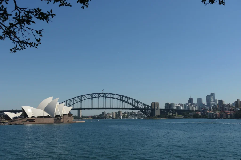 Blick über das Wasser auf das Sydney Opera House und die Harbour Bridge an einem sonnigen Tag, eingerahmt von Zweigen.