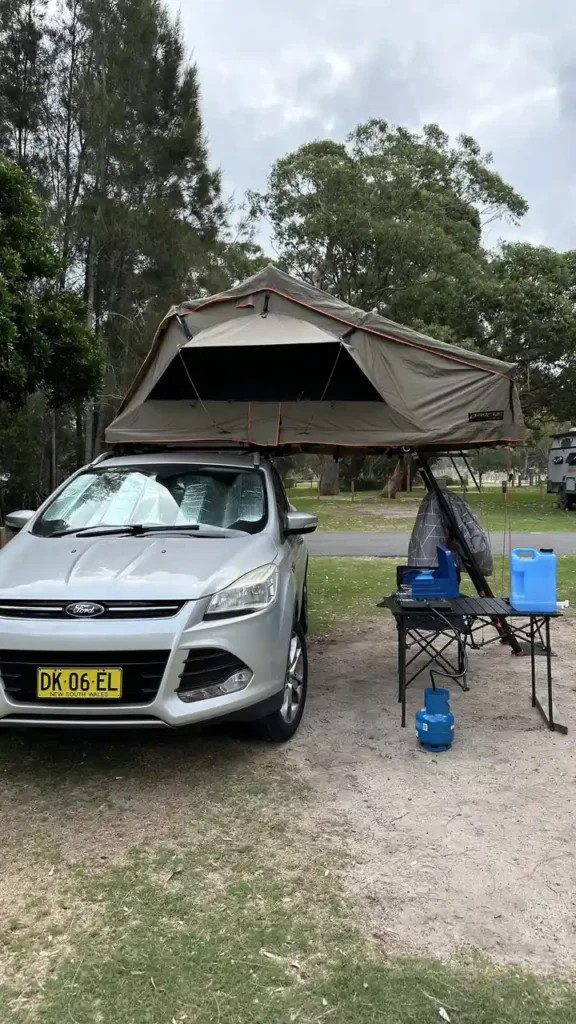 Ein silbernes Auto mit Dachzelt steht auf einer Wiese. Daneben Campingtisch, blaue Wasserkanister und Bäume wie beim Zelten.