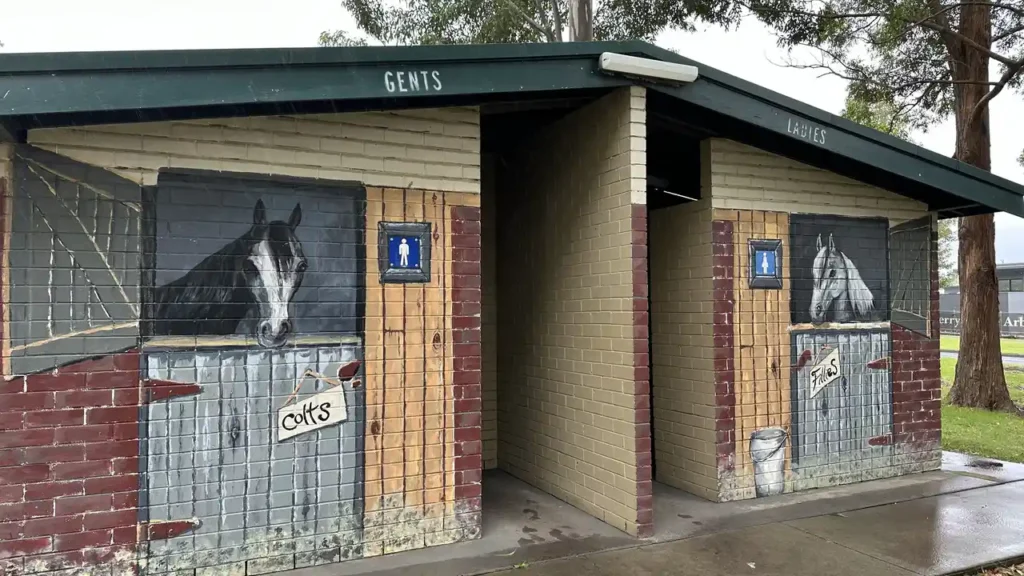 Öffentliche Toiletten mit getrennten Eingängen für Damen und Herren, bemalt mit Pferdemotiven; draußen nasser Boden und Bäume.