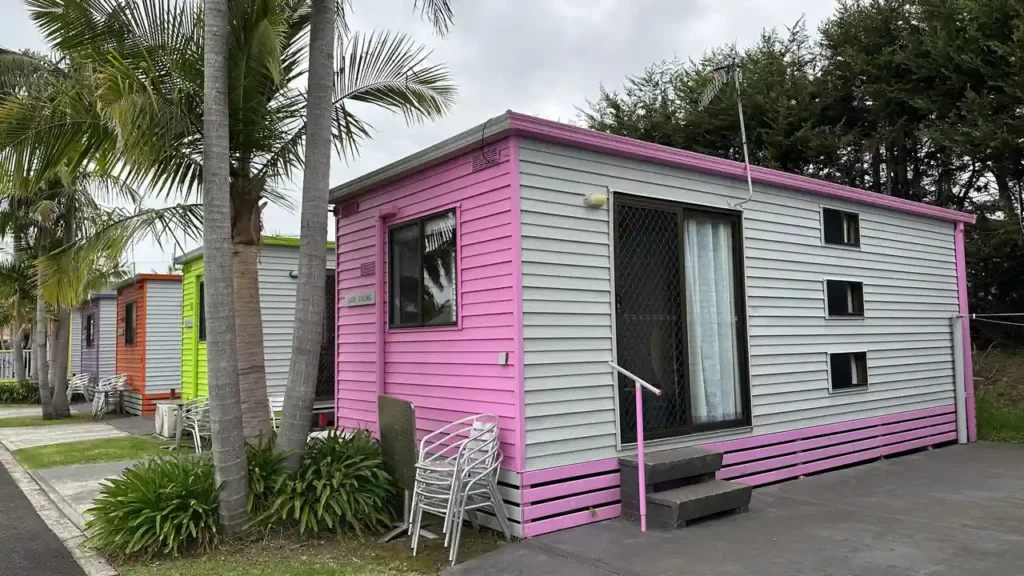 Ein kleines Haus mit grauer und pinker Fassade, Stufen zur Glastür, gestapelte Stühle bei einer Pflanze, Palmen und Wolken.