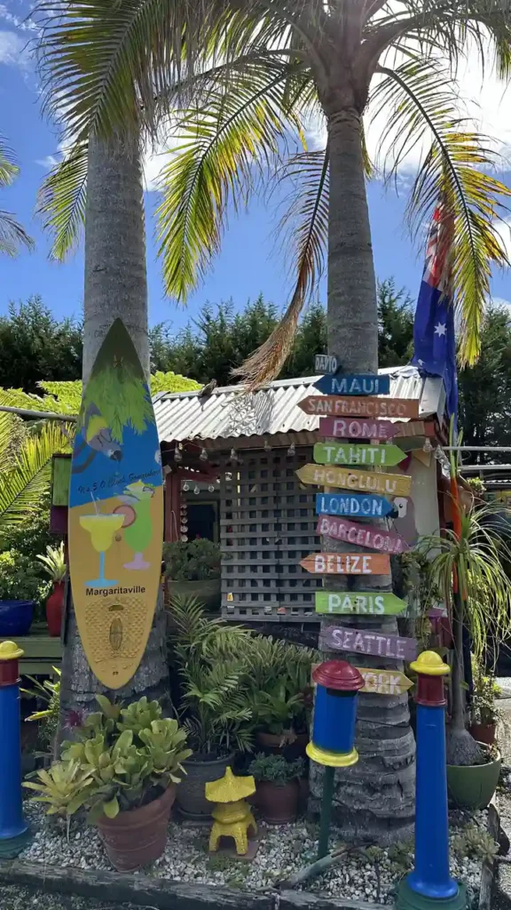 Bunte Tropenstimmung mit Palmen, Margaritaville-Surfbrett und Wegweisern nach Maui, Rom und Paris; im Hintergrund Hütte und Pflanzen.