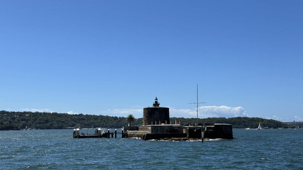 Ein kleiner Leuchtturm mit grünem Dach steht auf einer Felseninsel im blauen Meer, dahinter Bäume und ein Segelboot bei Sonnenschein.