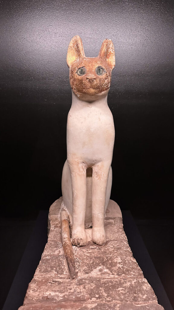 Eine altägyptische Katzenstatue mit rotbraunem Kopf und weißem Körper sitzt auf einem Stein, früher in Australien ausgestellt.