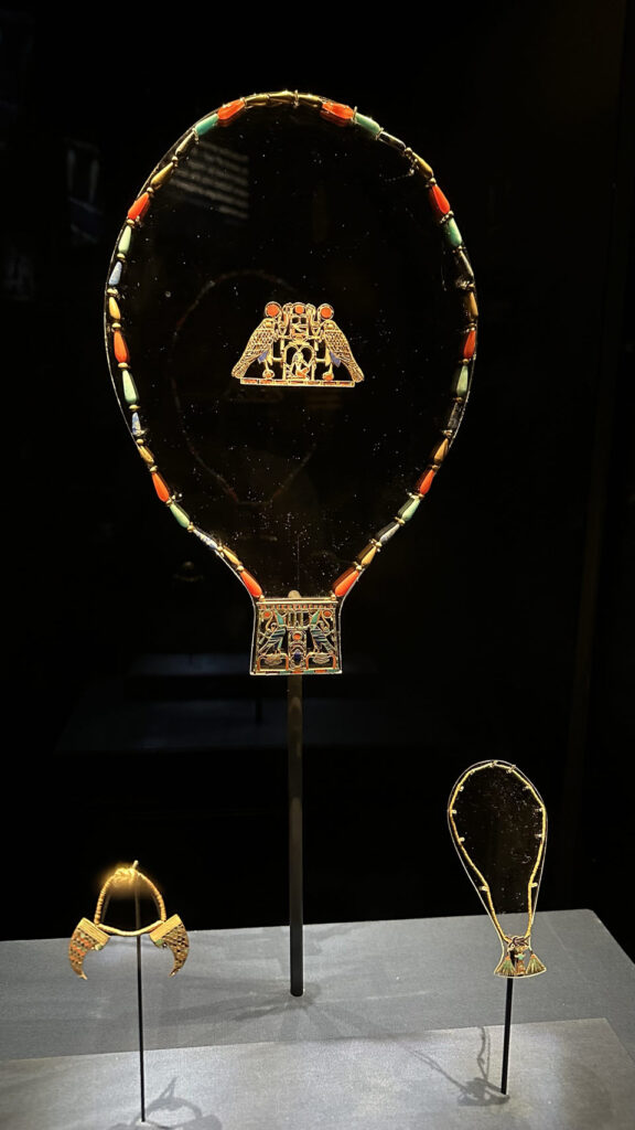 Drei antike ägyptische Schmuckstücke in einer Vitrine, darunter ein rundes, goldverziertes Stück mit Türkis und Karneol.