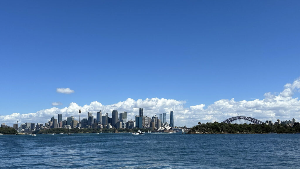 Blick über das Wasser auf Sydneys Skyline mit hohen Gebäuden, Opernhaus und Harbour Bridge unter blauem Himmel mit Wolken.