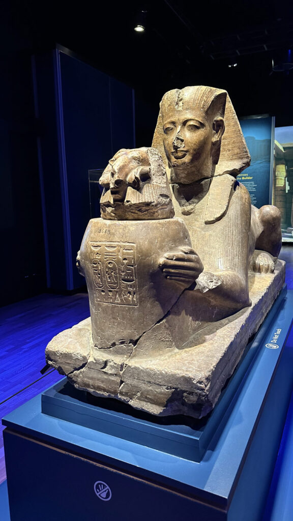 In einem australischen Museum steht eine steinerne Sphinx mit Pharaokopf, die eine kleinere, mit Hieroglyphen verzierte Sphinx hält.