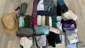 Zusammengerollte Kleidung, Badeanzüge und ein Strohhut auf dem Boden zum Probepacken – perfekte Reisevorbereitung vorm Rucksackpacken.