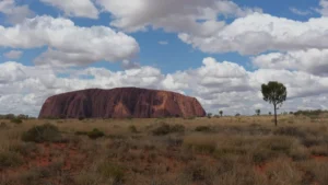 Uluru erhebt sich im Red Centre Australiens unter einem teilweise bewölkten Himmel inmitten trockenen Grases.